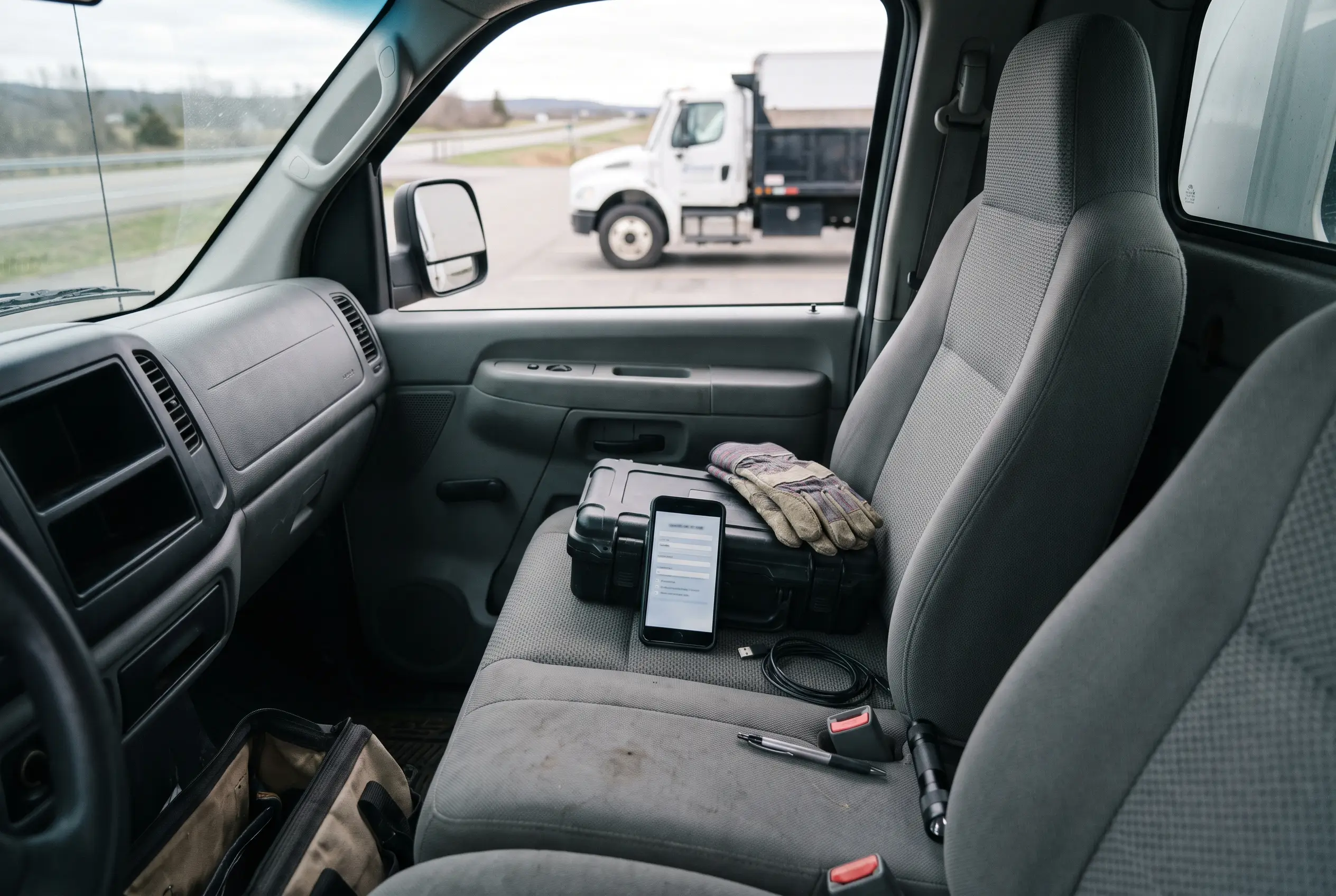 Truck cab seat holding a hard case, work gloves, a phone showing a form, a charging cable, and a pen, with a dump truck visible through the window