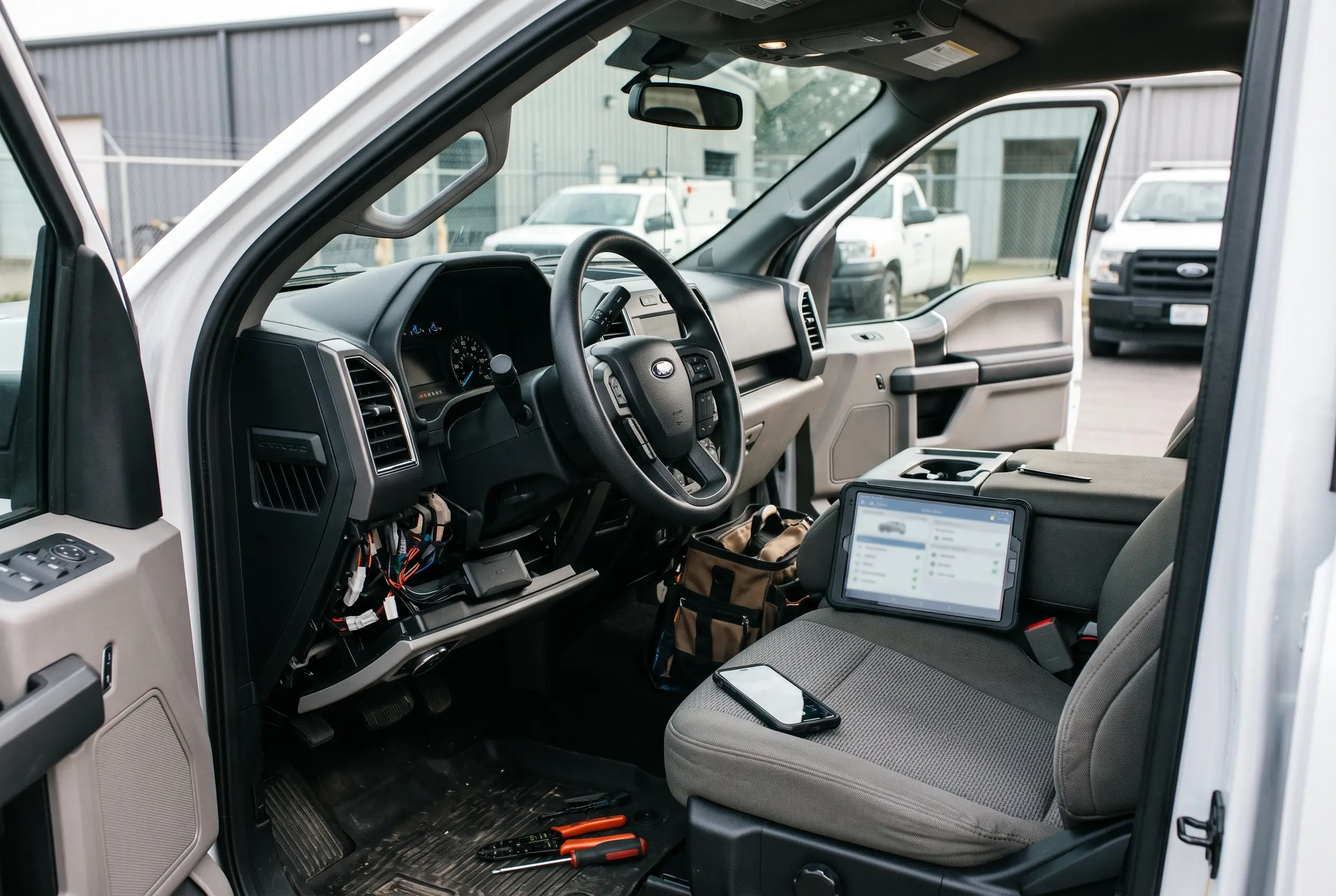 Pickup truck cab with the lower dash open and wiring exposed, a tablet and a phone on the seat, a tool bag, and hand tools on the floor mat